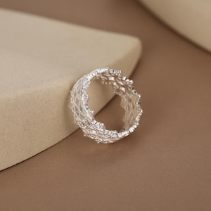 May include: A silver ring featuring an intricate, openwork lattice design with a textured, jagged edge. The metal has a bright, reflective finish, and the band is constructed from a delicate, interconnected web of small, metallic shapes.