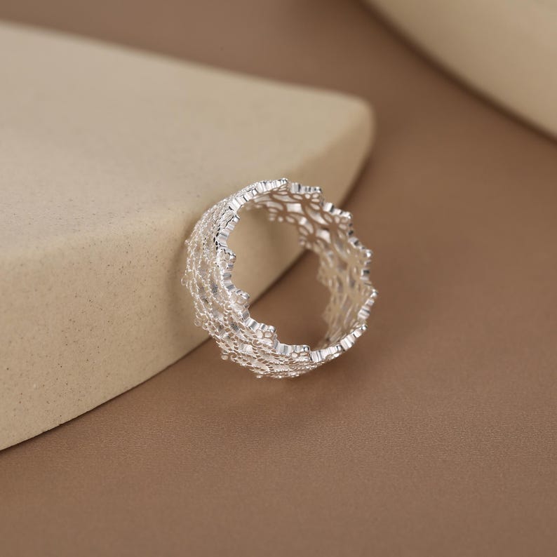 May include: A silver ring featuring an intricate, openwork lattice design with a textured, jagged edge. The metal has a bright, reflective finish, and the band is constructed from a delicate, interconnected web of small, metallic shapes.