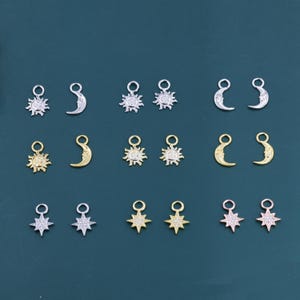 Op de afbeelding: Een verzameling kleine hemelse bedels in zilver, goud en roségoud tinten. De bedels omvatten zonnen, manen en sterren, elk met een kleine lus voor bevestiging. De bedels worden gepresenteerd op een donkerblauwe achtergrond.