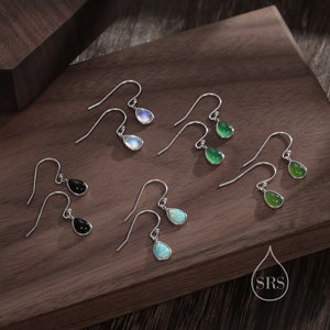 Op de afbeelding: Een collectie druppelvormige oorbellen in verschillende kleuren, waaronder zwart, groen, wit en opaal. De oorbellen worden gepresenteerd op een houten oppervlak. Elke oorbel heeft een zilveren haak.