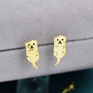 May include: A pair of gold-tone earrings featuring a cute otter design. The otters are depicted in a sitting position with their paws clasped together.