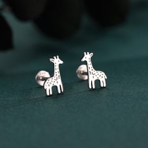 May include: A pair of silver giraffe stud earrings. The earrings have a polished finish and feature detailed spots. The giraffe earrings are set against a dark green background.