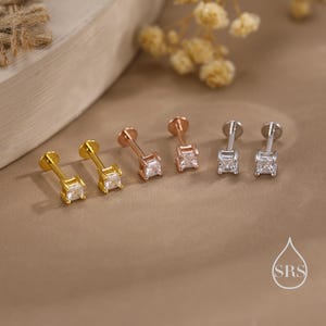 May include: Six stud earrings in gold, rose gold, and silver, each with a square, clear gemstone. The earrings are presented on a neutral surface, with a soft-focus background of dried flowers and a wooden element.