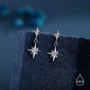 May include: Silver-toned starburst earrings with a dangling design. Each earring features a small star at the top and a larger star with a central clear stone. The earrings are displayed on a dark blue textured surface.