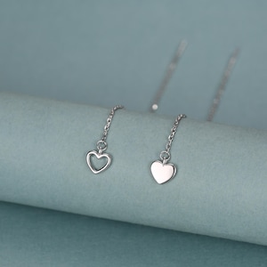 May include: A pair of silver-toned threader earrings featuring delicate chain links. One earring displays an open heart outline charm, while the other earring features a solid, smooth heart-shaped charm, both suspended from thin, polished metal chains.
