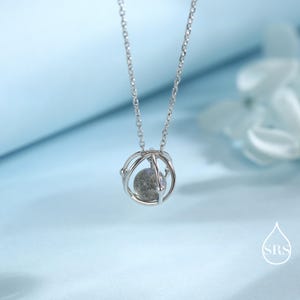 May include: A silver necklace featuring a pendant with a spherical, speckled stone encased in a cage-like design. The chain is delicate, and the pendant hangs against a soft blue background. The necklace is a piece of jewelry.