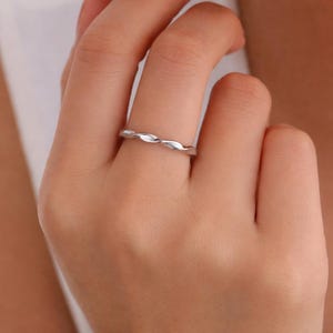 Può includere: Un sottile anello color argento lucido caratterizzato da una texture continua ritorta o simile a una corda lungo l'intera fascia. Il metallo presenta una finitura liscia e riflettente che cattura la luce, creando un motivo sottile e ripetitivo.