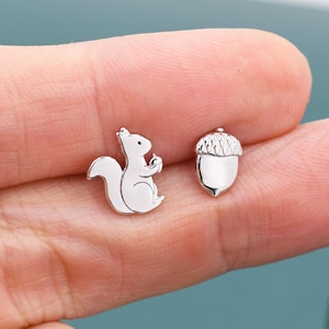 May include: A pair of silver-colored stud earrings. One earring is shaped like a squirrel holding an acorn, and the other is an acorn. The earrings are small and detailed, with a shiny finish.