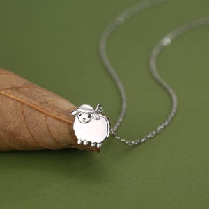 May include: A silver-toned necklace featuring a small, stylized sheep pendant with a smooth, polished finish. The sheep has simple, engraved facial features including closed eyes and a small mouth. The pendant hangs from a delicate silver chain.