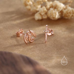 Peut inclure: Boucles d'oreilles clous en or rose. Une boucle d'oreille pr&eacute;sente un motif de dragon, l'autre une &eacute;p&eacute;e. Les boucles d'oreilles sont pr&eacute;sent&eacute;es sur une surface marron avec des fleurs s&eacute;ch&eacute;es en arri&egrave;re-plan. Les boucles d'oreilles sont petites et d&eacute;licates.