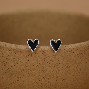 Puede incluir: Un par de pendientes de botón en forma de corazón. Cada pendiente presenta un corazón de esmalte negro engastado en un metal plateado. Los pendientes se muestran sobre una superficie marrón claro neutra.
