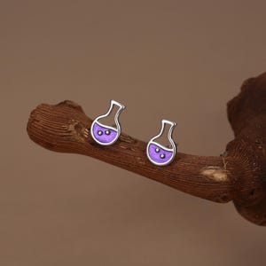 May include: A pair of silver-colored stud earrings shaped like laboratory flasks. Each flask has a purple liquid and two small bubbles inside. The earrings are displayed on a brown, textured wooden branch against a brown background.