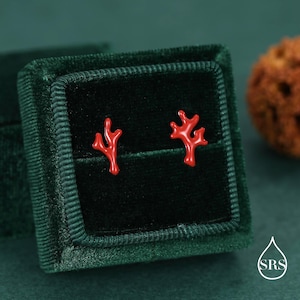 Puede incluir: Un par de pendientes de botón con forma de coral rojo brillante, presentados en una caja de joyería de terciopelo verde oscuro. Los pendientes son el punto focal, con un diseño detallado de coral. La caja está abierta.