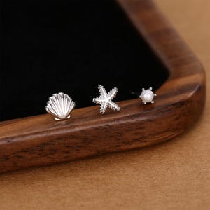 Puede incluir: Tres pendientes de botón plateados con diseños de temática marina. Los pendientes incluyen una concha, una estrella de mar y un pendiente con forma de perla. Los pendientes se muestran sobre una superficie de madera con un fondo negro.