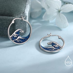 Puede incluir: Un par de pendientes de aro de plata con un dise&ntilde;o de ola en esmalte azul y blanco. Cada pendiente circular muestra un motivo de ola oce&aacute;nica estilizada con coloraci&oacute;n azul intenso y detalles de espuma blanca en el dise&ntilde;o.