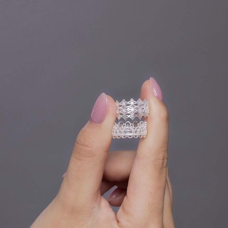 May include: A silver ring featuring an intricate, openwork lace-like pattern that resembles a crown. The band has a delicate, repeating filigree design with small, circular cutouts throughout the metal, creating a light and airy appearance.