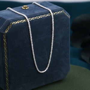 Puede incluir: Un collar de cadena de plata con un dise&ntilde;o delicado y brillante. El collar se muestra en una caja de joyer&iacute;a de terciopelo azul.