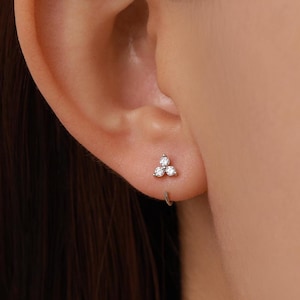 May include: A silver-toned stud earring featuring a cluster of three small, clear, round-cut stones arranged in a triangular formation. The earring is secured with a thin, curved metal wire that loops behind the earlobe, creating a delicate look.
