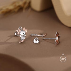 Peut inclure: Une paire de boucles d'oreilles en argent avec un motif d'axolotl souriant. Une boucle d'oreille a un axolotl en or rose, tandis que l'autre boucle d'oreille est un simple clou en argent.