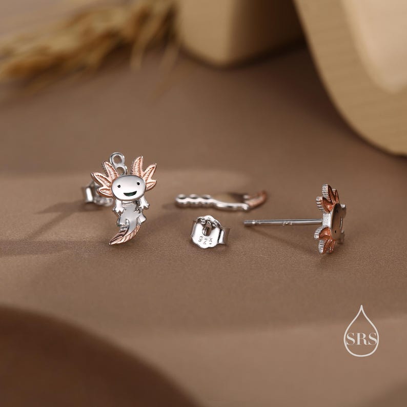 Peut inclure: Une paire de boucles d'oreilles en argent avec un motif d'axolotl souriant. Une boucle d'oreille a un axolotl en or rose, tandis que l'autre boucle d'oreille est un simple clou en argent.