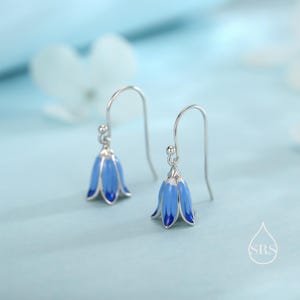 May include: Silver-coloured earrings with blue bellflower pendants and dark blue edges. The earrings have a hook fastening and are set against a light blue backdrop. The floral design adds a touch of nature-inspired elegance.