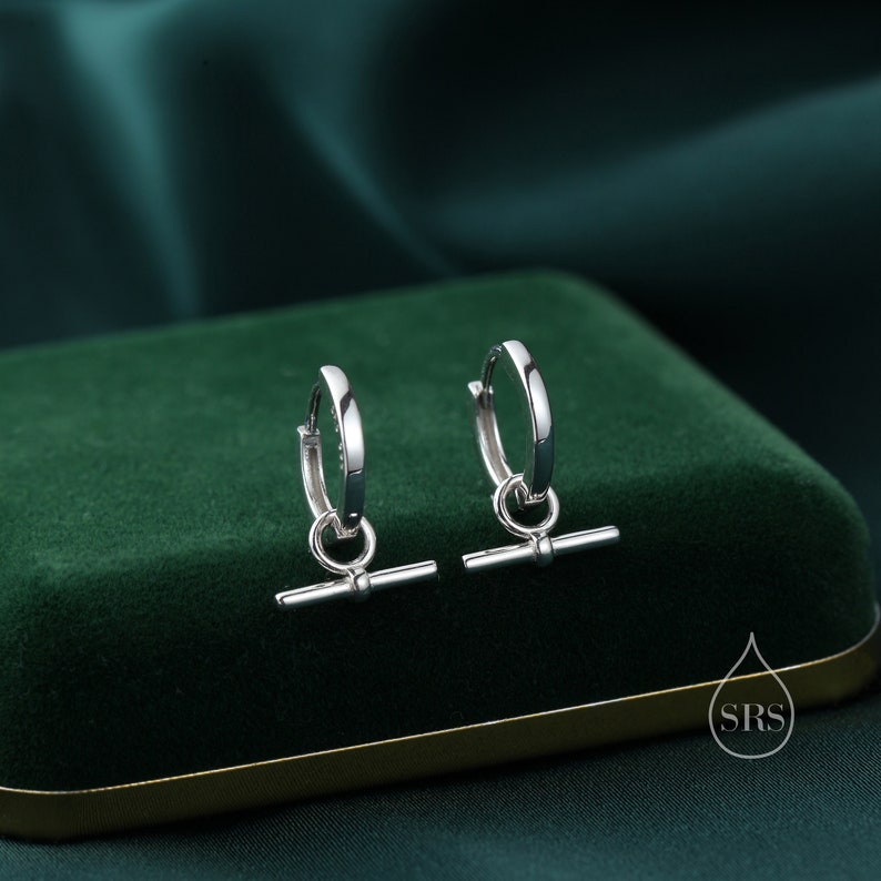 May include: A pair of silver hoop earrings with a small bar hanging from each hoop. The earrings are on a green velvet surface.