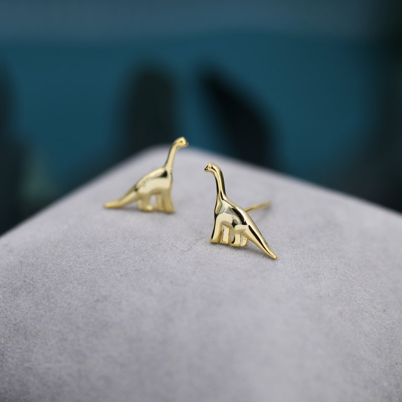 May include: A pair of gold-toned dinosaur stud earrings. The earrings are small and delicate, and they feature a realistic dinosaur design.