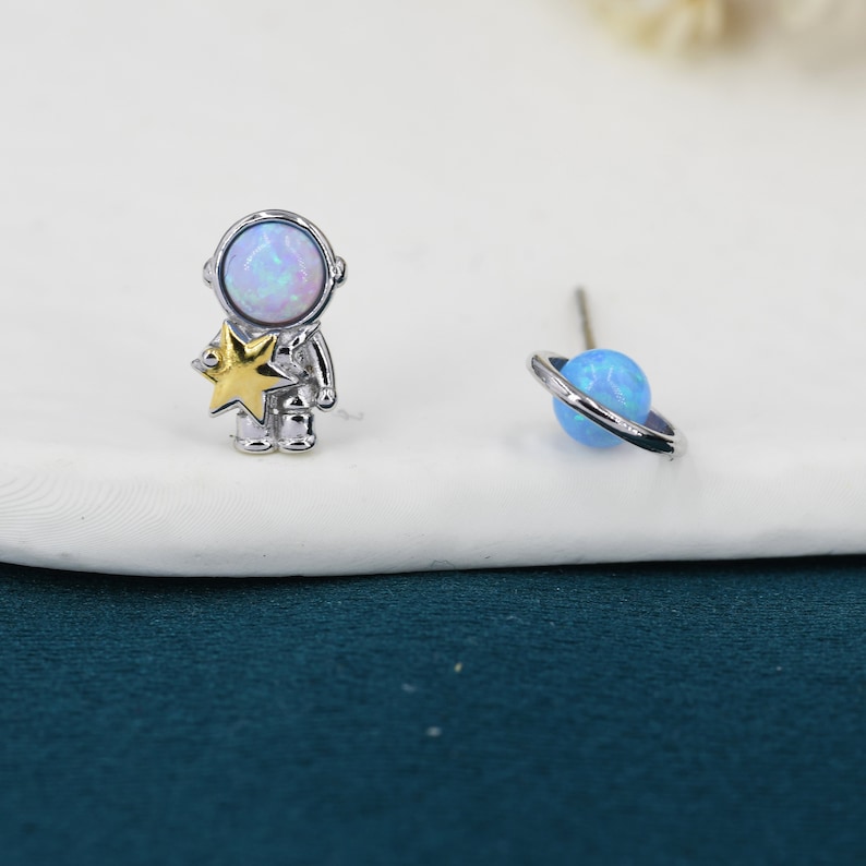 May include: A pair of silver-toned stud earrings with an outer space theme. One earring features an astronaut holding a gold star, and the other depicts a blue planet with a silver ring around it. The astronaut's helmet and the planet have a pearlescent finish.