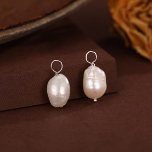 May include: Two white, baroque pearl earrings with silver-colored hoops. The pearls have a lustrous sheen and irregular shapes. The earrings are displayed on a brown surface.