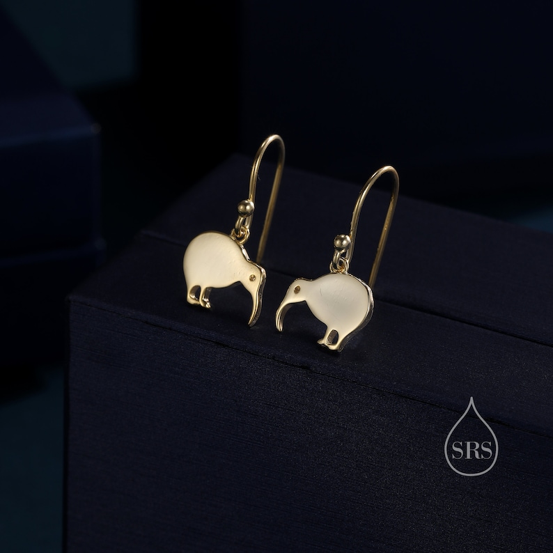 May include: A pair of gold earrings featuring a kiwi bird design. The earrings are made of metal and have a white enamel finish.