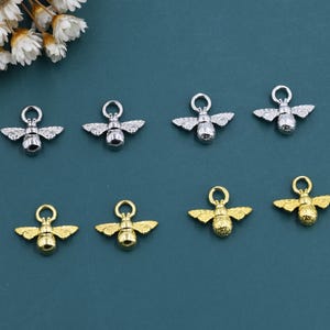 May include: Eight bee-shaped charms in silver and gold tones. The charms have detailed wings and a loop at the top for attaching to jewelry. The background is a teal color, and there are white flowers in the upper left corner.