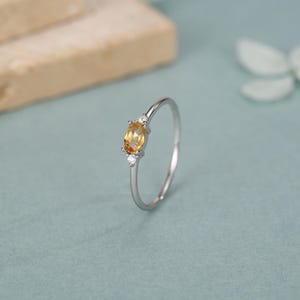 Könnte beinhalten: Ein dünner Silberring mit einem zentralen, oval geschliffenen gelben Citrin, der von zwei kleinen, klaren, rund geschliffenen Akzentsteinen flankiert wird. Der schmale, polierte Metallring hält die drei Steine in einer Krappenfassung.