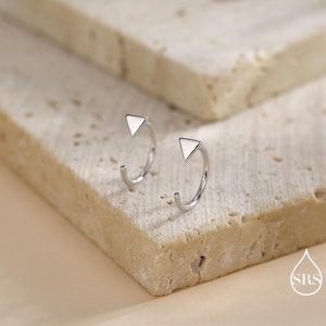 Può includere: Un paio di orecchini a cerchio color argento con un design a triangolo bianco. Gli orecchini aperti sono appoggiati su una superficie chiara e strutturata. Gli orecchini hanno uno stile moderno e minimalista.
