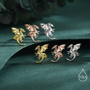May include: Six dragon-shaped stud earrings in gold, rose gold, and silver. The earrings feature detailed wings and a curved tail. The earrings are displayed on a dark green surface.
