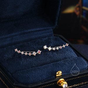 May include: A pair of silver ear cuffs with a curved design. Each cuff features a row of small, clear and red gemstones.