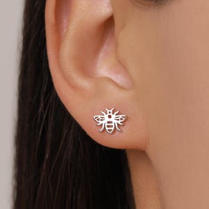 May include: A silver-colored bee stud earring is shown on an ear. The earring is a detailed cutout of a bee, showcasing its wings, body segments, and antennae. The earring is small and delicate, suitable for everyday wear.
