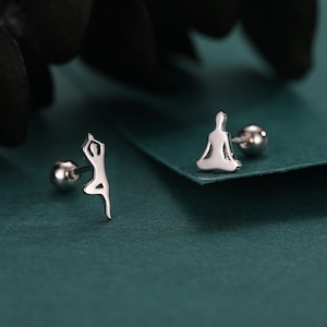 May include: Two silver stud earrings. One earring features a figure in a yoga pose, the other depicts a person in a seated meditation position. The earrings are set against a dark teal background.