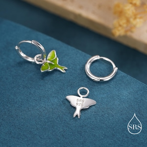 May include: Silver-toned earrings with a green and white design, featuring a small charm with the text "SRS 925". The earrings are set against a blue background. The earrings are a hoop style.