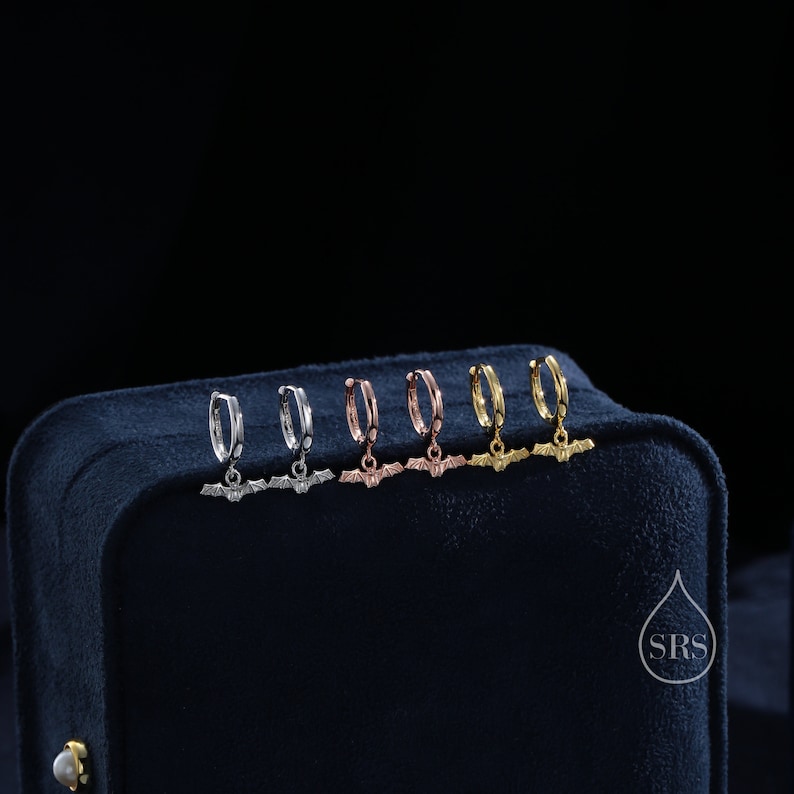 May include: Five pairs of hoop earrings with a bat charm hanging from each hoop. The hoops are silver, rose gold, and gold. The earrings are displayed on a black velvet box.