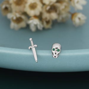 Op de afbeelding: Zilverkleurige oorstekers. Eén oorbel is een kleine dolk, de andere is een schedel met groene edelsteenogen. De oorbellen worden gepresenteerd op een lichtblauwe ondergrond met een vage bloemenachtergrond.