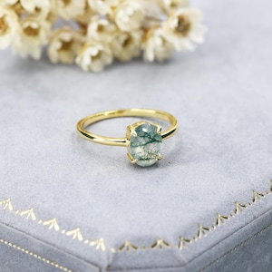 May include: A gold ring with a green moss agate oval gemstone. The ring is on a gray velvet background.