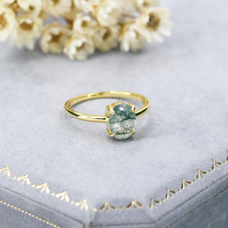 May include: A gold ring with a green moss agate oval gemstone. The ring is on a gray velvet background.