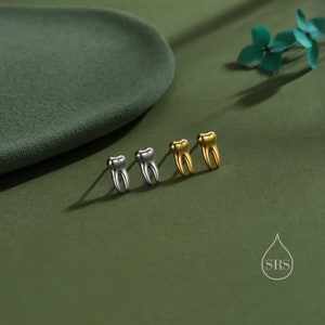 May include: Two pairs of stud earrings shaped like human teeth. One pair is finished in a polished silver tone, and the other pair is finished in a polished gold tone. Each earring features a post back. A small logo with the letters SRS is visible.