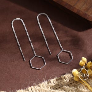 Puede incluir: Un par de pendientes de cadena plateada con dijes hexagonales. Los dijes son huecos y tienen una forma geométrica.