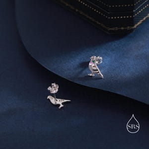 May include: A pair of silver bird earrings with green and purple enamel accents. The earrings are on a blue background.