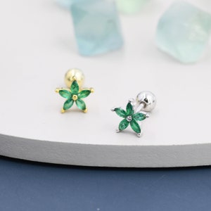 May include: Two delicate flower-shaped earrings with green gemstones. The earrings are on a white surface. One earring has a gold post and the other has a silver post.