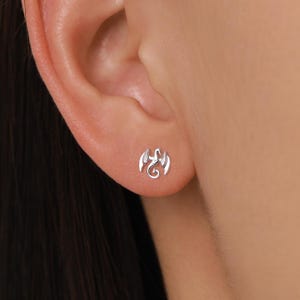 May include: A small, silver-toned stud earring shaped like a dragon with its wings spread upward. The mythical creature design features a curved tail and detailed wing structures, positioned centrally on the earlobe as a single piece of jewelry.