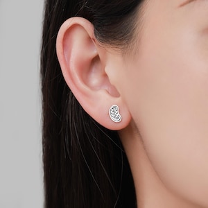 May include: A silver-colored stud earring in the shape of a kidney is shown on an ear. The earring has a detailed, textured design. The background is a blurred view of a person's skin and hair.