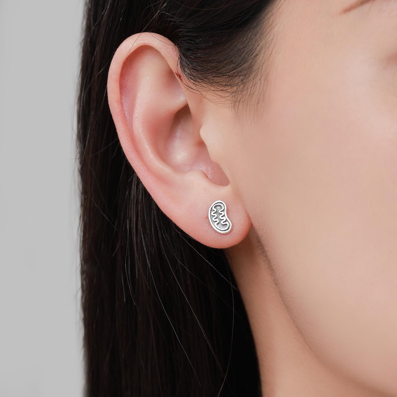 May include: A silver-colored stud earring in the shape of a kidney is shown on an ear. The earring has a detailed, textured design. The background is a blurred view of a person's skin and hair.