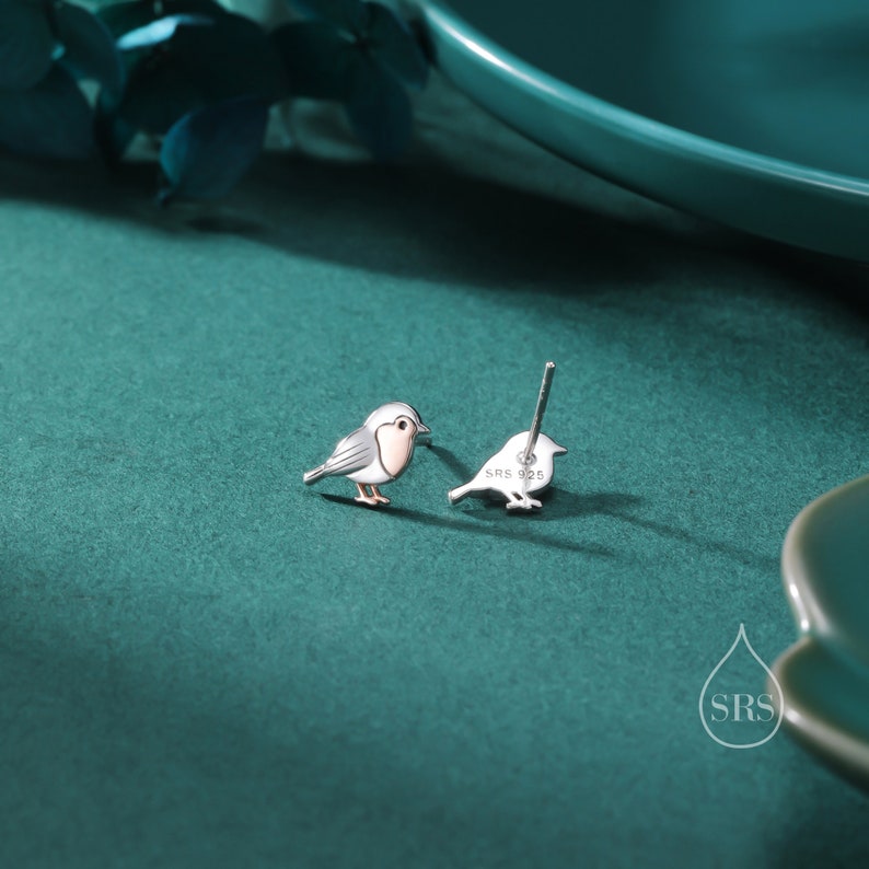 K&ouml;nnte beinhalten: Ein Paar silberne Ohrringe mit einem kleinen, detaillierten Rotkehlchen-Design. Das Rotkehlchen ist in hellgrau mit einer rosa Brust dargestellt. Die Ohrringe sind auf der R&uuml;ckseite mit "SRS 925" gekennzeichnet.